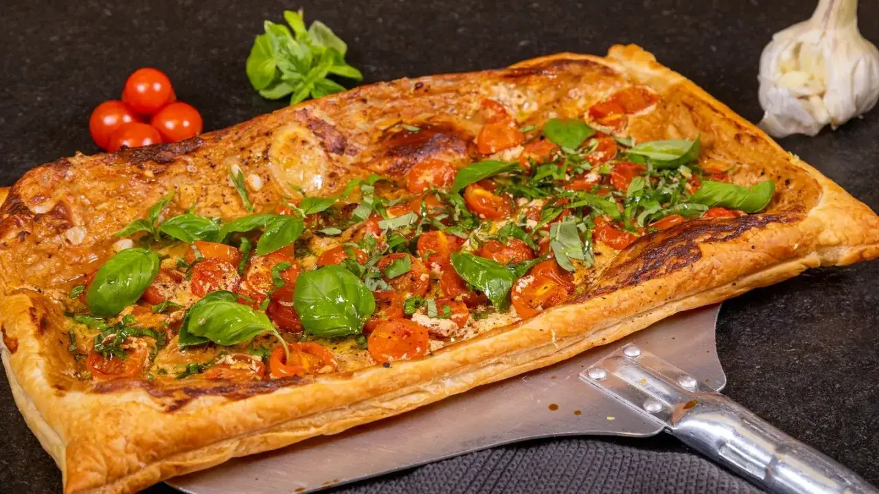 Herzhafte tarte mit tomaten und frischem Basilikum, goldbraun gebacken. Daneben ein Knoblauch und Kirschtomaten.