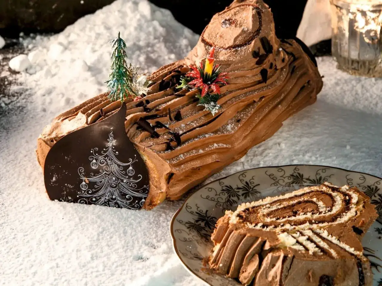 Festliche B&ucirc;che de No&euml;l, dekoriert mit Schokost&uuml;ckchen und einer Kerze, liegt im Schnee. Ein St&uuml;ck auf einem Teller zeigt die Spiralen.