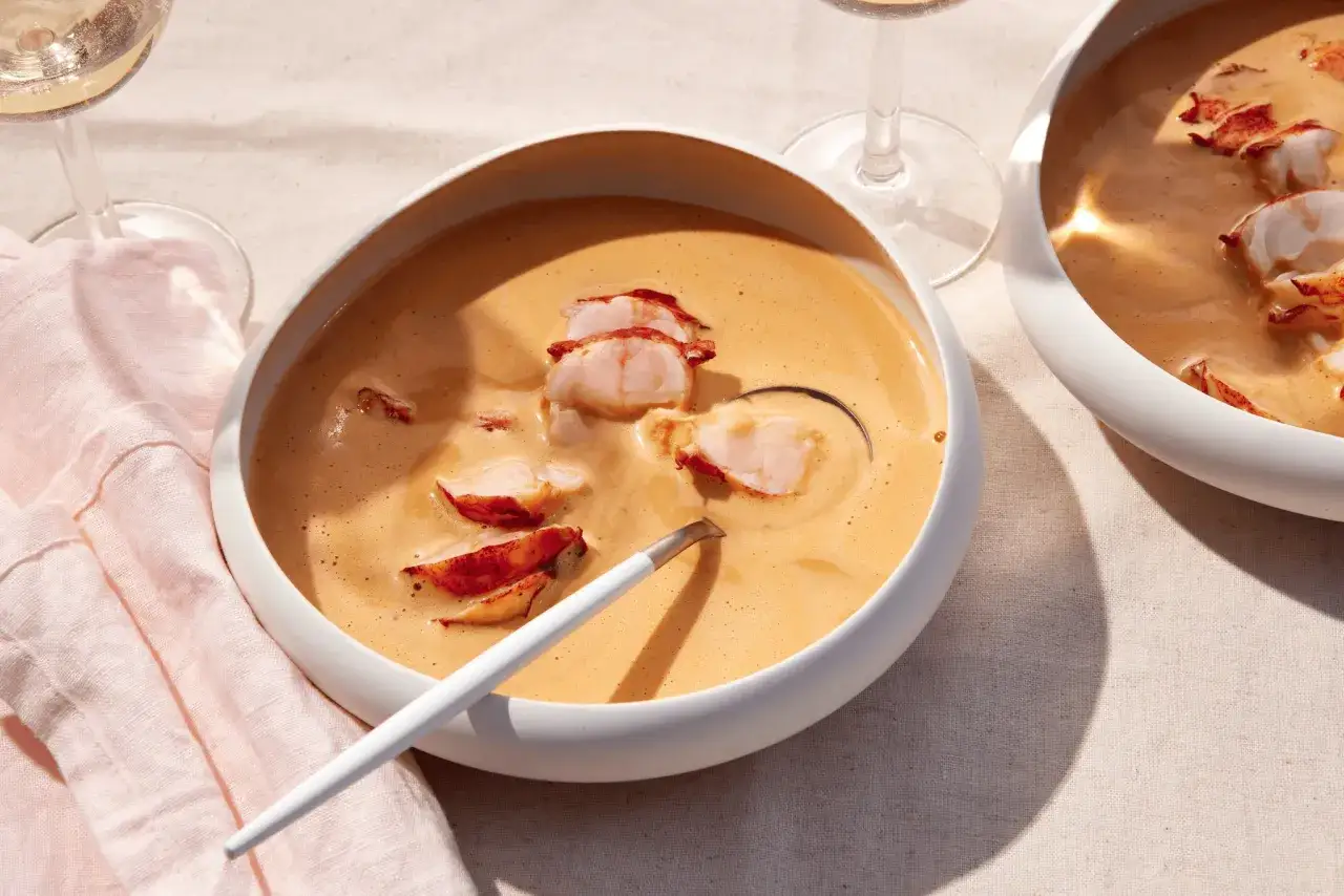 Cremige Hummerbisque mit zarten Hummerst&uuml;cken in einer wei&szlig;en Sch&uuml;ssel, daneben ein Glas Wei&szlig;wein.