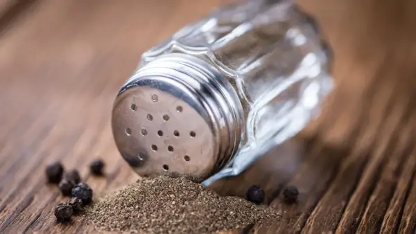 Ein Glasstreuer mit Metallaufsatz liegt auf einem Holztisch. Gemahlener Pfeffer und ganze Körner sind daneben verstreut. So kann man den Pfeffer neutralisieren.