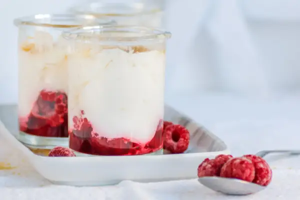 Zwei Gläser mit cremigem Dessert und Himbeeren. Unten leuchtet ein intensiver Himbeercoulis, gekrönt von luftiger Sahne. Frische Himbeeren zieren die Gläser und eine Löffelspitze.