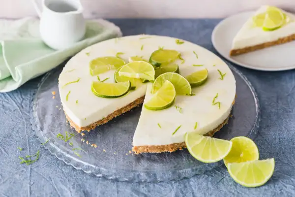 Erfrischender limetten cheesecake ohne backen, dekoriert mit frischen Limettenspalten und Zesten. Ein Stück ist bereits angeschnitten.