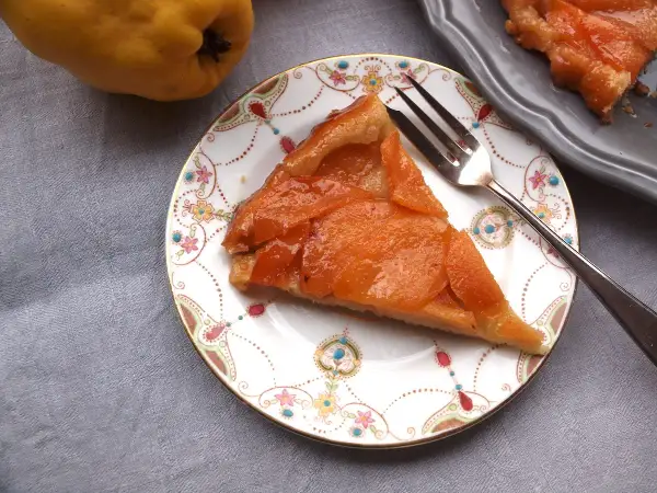 Ein Stück Quitten Tarte Tatin auf einem Teller mit Blumenmuster. Daneben eine ganze Quitte und ein weiteres Stück Tarte.