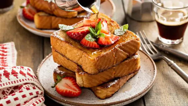 Ein Stapel goldbrauner Pain Perdu, garniert mit Erdbeeren und Minze, wird mit Sirup übergossen. Daneben ein Glas Kaffee.