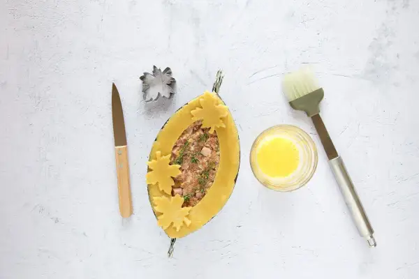 Ein ovaler Teigmantel, gefüllt mit Hackfleisch und Kräutern, wird mit Blätter-Ausstechern verziert. Daneben liegen ein Messer, ein Ausstecher, Eigelb und ein Pinsel für das perfekte Pate en croute.