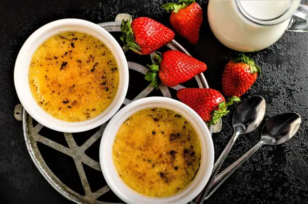 Zwei cremige Crème brûlée mit karamellisierter Zuckerkruste, garniert mit frischen Erdbeeren und einem Milchkännchen.