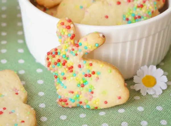 Bunte Osterkekse zum Ausstechen in Hasenform, verziert mit bunten Streuseln, liegen auf einem grünen Tupfentuch neben einer weißen Schüssel.