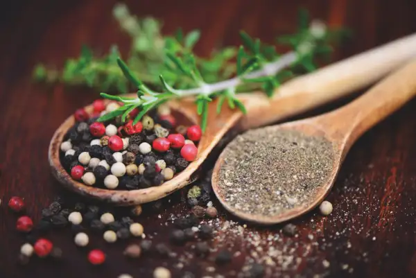 Zwei Holzlöffel mit Pfefferkörnern und gemahlenem Pfeffer. Ein Zweig Rosmarin liegt darüber. Ist Pfeffer gesund? Ja, er würzt und ist gut für dich!