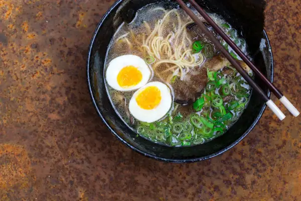 Schüssel mit dampfendem Ramen, zwei halbierten Eiern, Nudeln, Fleisch und Frühlingszwiebeln. Perfekt, um Ramen selbst zu machen!